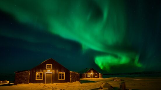 Bright green northern lights swirl in the night sky over two small lit cabins on a snow-covered ground.