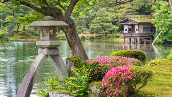 A stone lantern and pink azaleas overlook a calm pond with a traditional wooden tea house on stilts in the background.