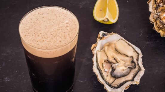 A glass of dark stout beer with foam next to an open raw oyster and a slice of lemon on a dark surface.