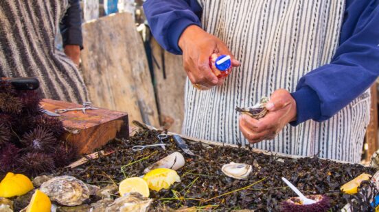 A person in a striped apron shucking a fresh oyster over a table of seafood and sliced lemons.