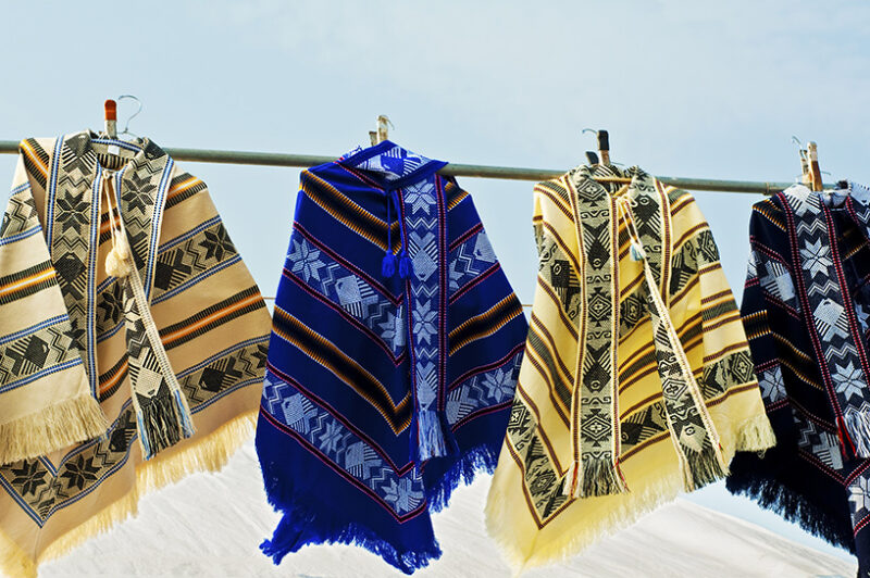 Four brightly colored, patterned ponchos with fringe hanging side-by-side on a metal rod outside. Luxury Argentina tours.