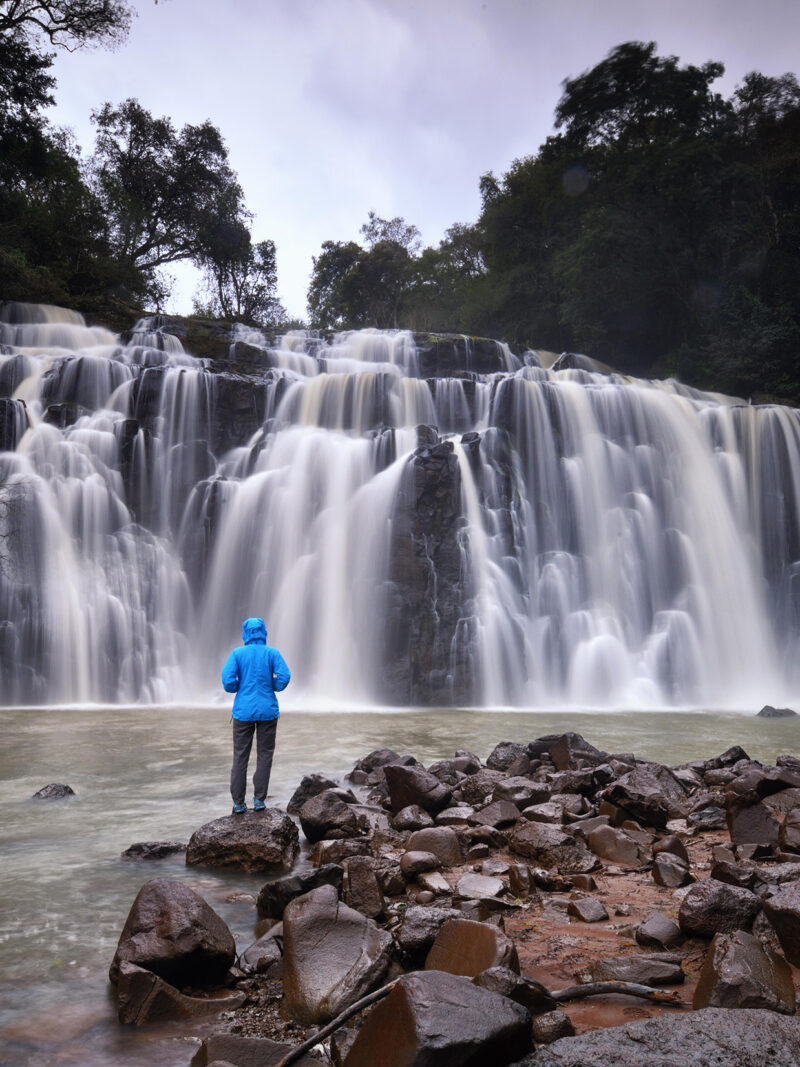A person in a blue jacket stands on rocks, facing a wide, multi-tiered waterfall on a cloudy day. Luxury Argentina tours.
