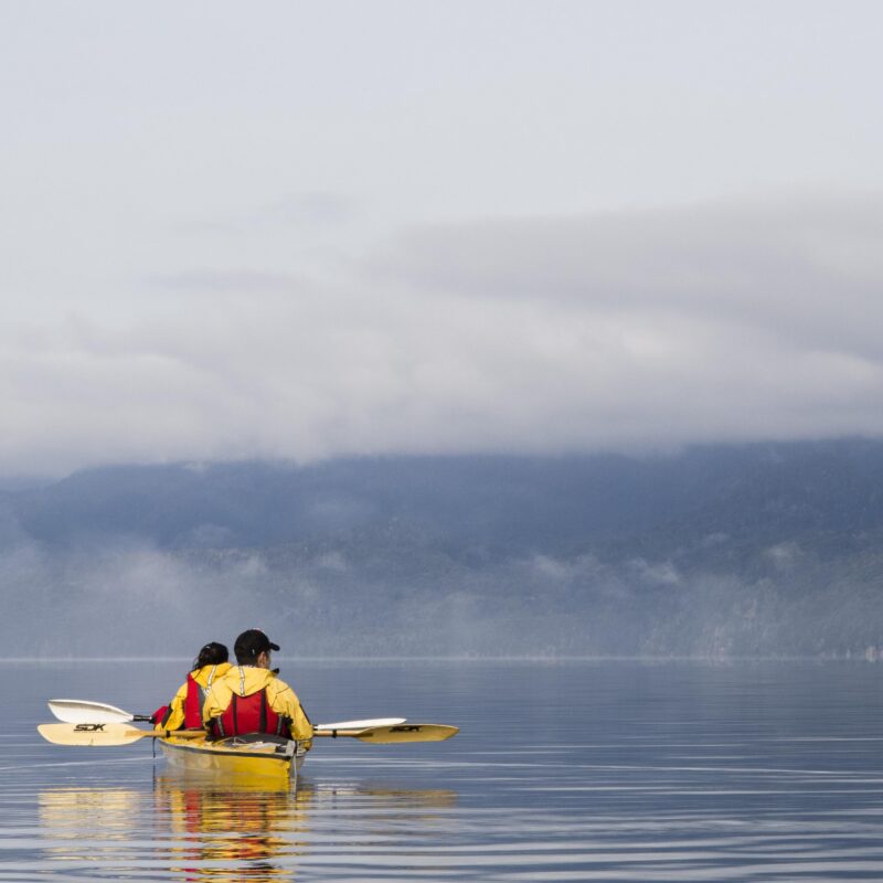 Two people in a yellow kayak paddle on a calm lake with foggy, tree-covered mountains in the distance. Luxury Argentina tours.