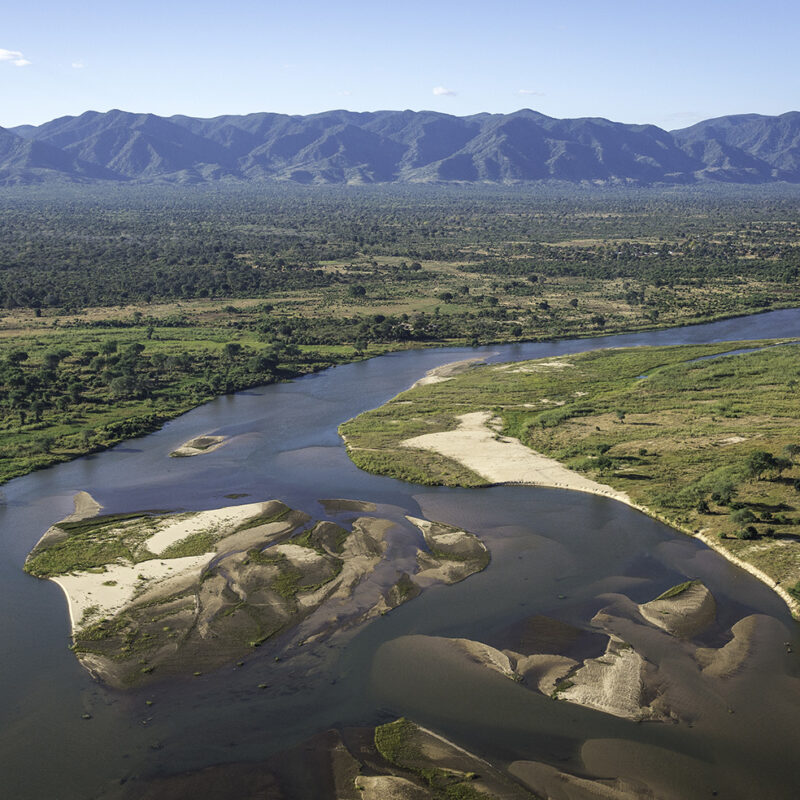Aerial view of a wide river with sandbanks running through a forested valley with a large range of mountains in the distance.