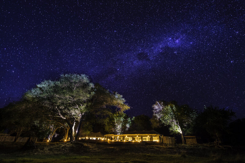 An illuminated luxury safari camp, visible under a spectacular dark night sky full of bright stars and the Milky Way.