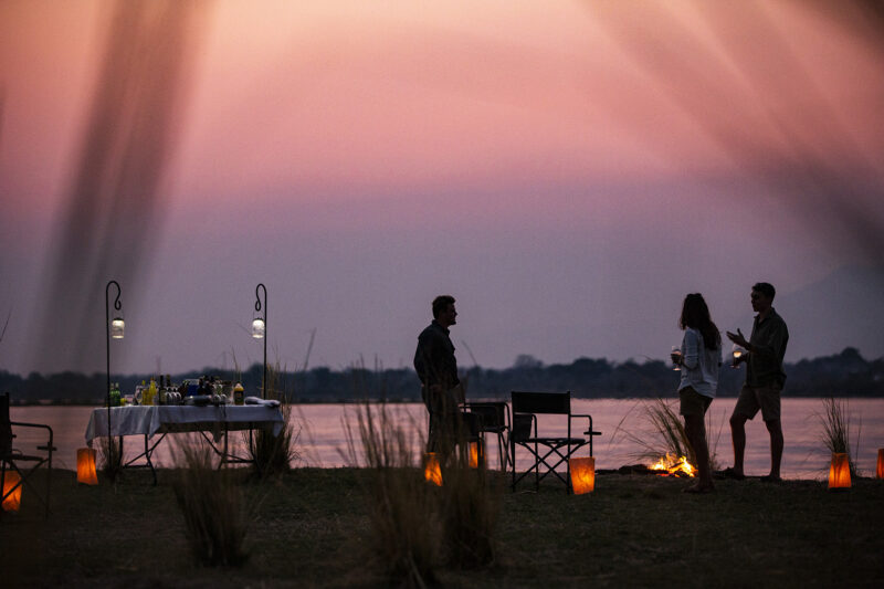 Silhouettes of three people standing by a river at sunset, with a bar table and lanterns lit on the grassy bank.