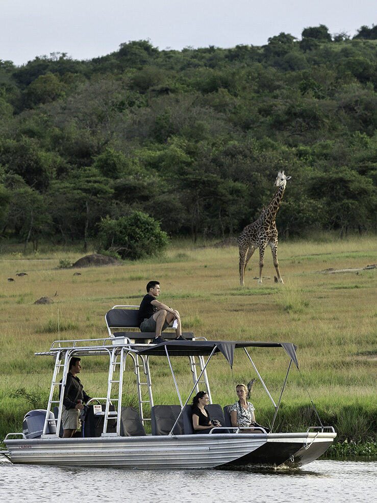 Tourists on a covered boat watching a giraffe on the grassy riverbank in the distance. Book luxury Rwanda safaris today.