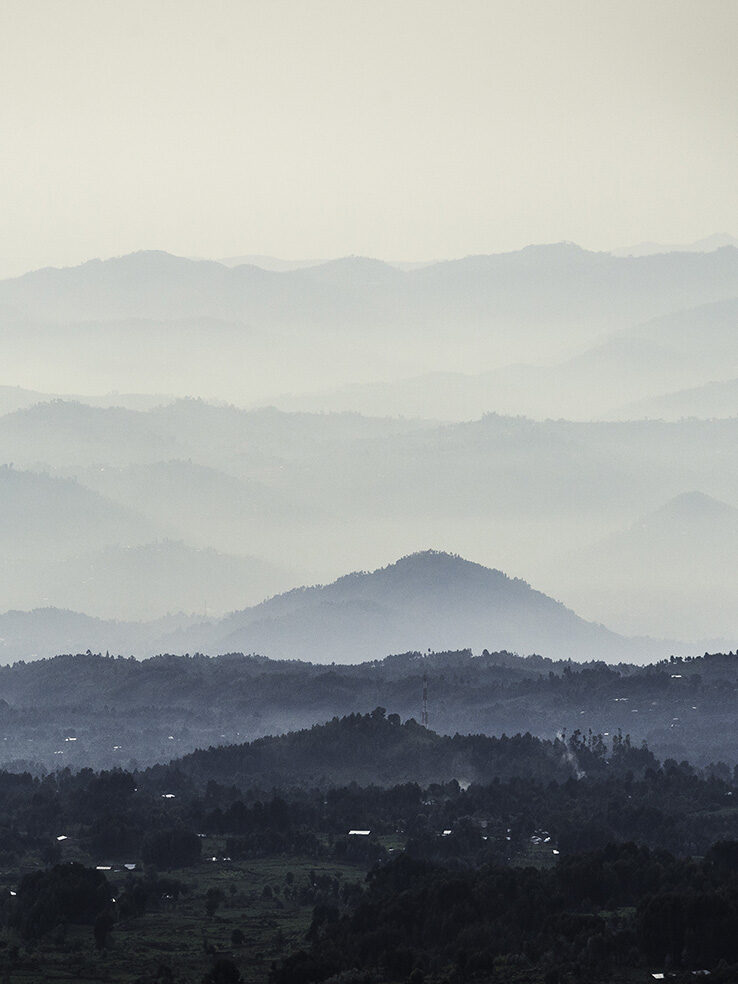 Wide view of tiered rolling hills and valleys covered in layers of blue and white mist. Enjoy the landscapes on luxury Rwanda vacations.
