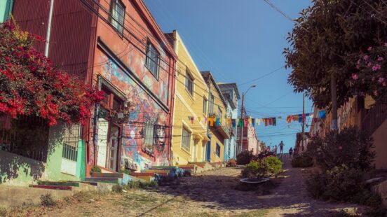 A steep, cobbled street in Valparaíso is lined with bright, colorful houses and red flowers on the left.