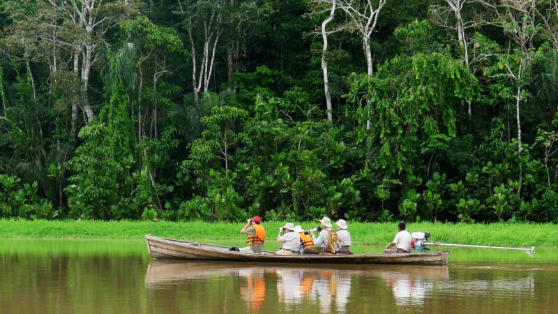 A group of five tourists and a guide are riding in a long, wooden motorized boat on a wide, calm brown river, backed by dense green jungle foliage in the Amazon during Luxury Colombia vacations.