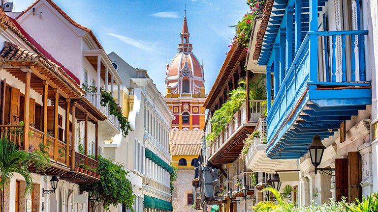 A street view of historic, colorful colonial buildings with balconies and lush plants, leading toward the distinct orange dome of a central tower in Cartagena, part of Luxury Colombia Trips.