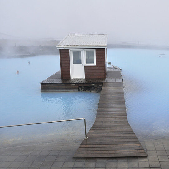 A small brown wooden cabin on a deck is surrounded by the milky blue water of the Blue Lagoon, promoting luxury Iceland tours.