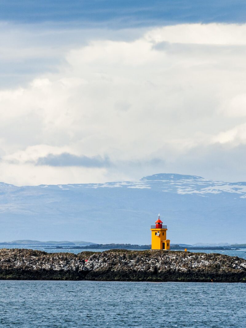 A small, bright yellow lighthouse stands on a dark rocky breakwater