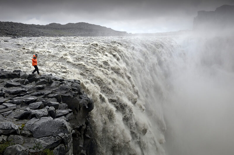 A person in an orange jacket stands on dark rocks at the edge of a massive, misty waterfall, perfect for luxury Iceland trips.