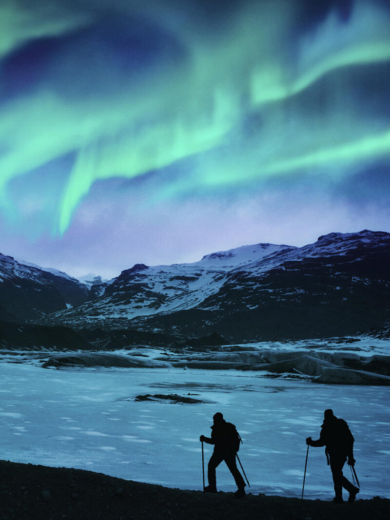 Two hikers stand silhouetted on a frozen glacier under the blue and green Northern Lights, highlighting luxury Iceland tours.