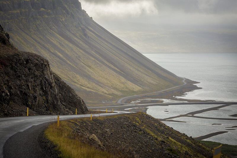 A winding road curves along a steep, grassy coastal mountainside next to water, perfect for luxury Iceland trips.