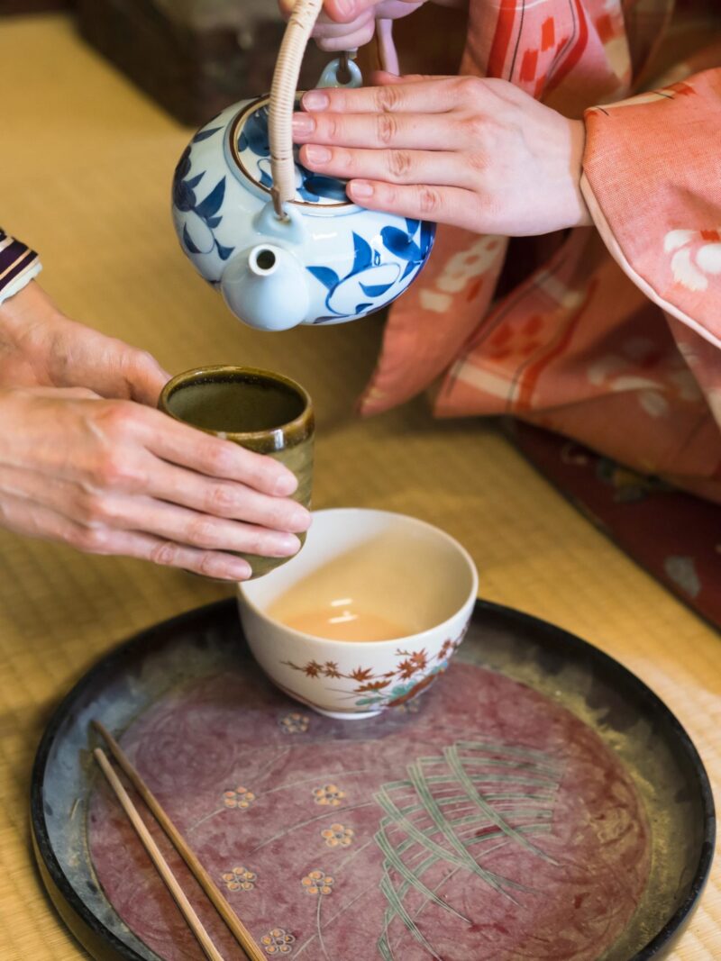 Private Japan Luxury Tours - detail of two women in traditional kimono, kneeling on tatami preparing, pouring a cup of tea which is in the hands of one woman. They are in traditional Japanese old house on tatami.