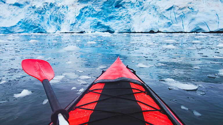 First-person view from a red kayak with a paddle facing a massive blue and white iceberg wall in icy water.