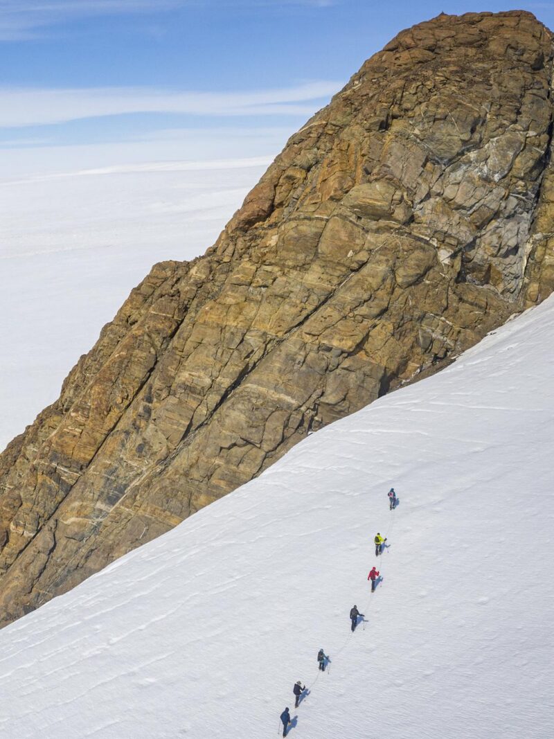 A line of climbers ascend a steep snow and ice slope next to a massive, brown, rocky mountain wall.
