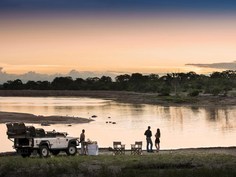 Luxury South African Tours - People stand by a calm lake at sunset next to a white safari vehicle and a table with white cloth.