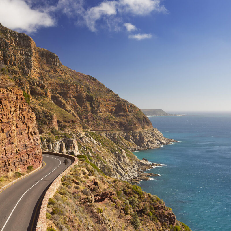 Luxury South African Tours - A high-angle view of a coastal road curving along steep mountains next to the blue sea.