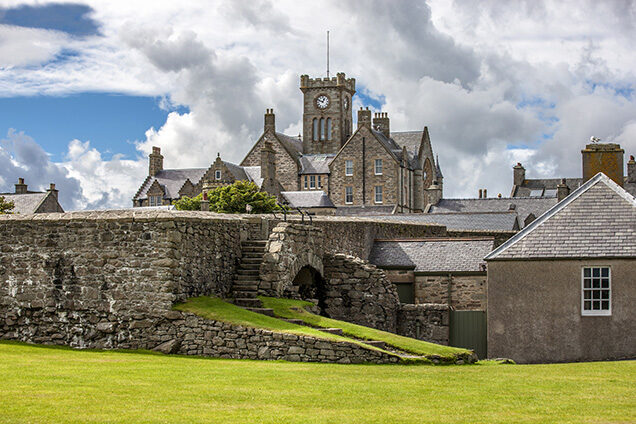 Stone building with a clock tower and a traditional stone boundary wall with stairs leading to an archway.
