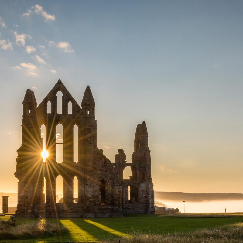 Ruins of Whitby Abbey silhouetted against the bright sun on the horizon, casting a sunburst through the arches.