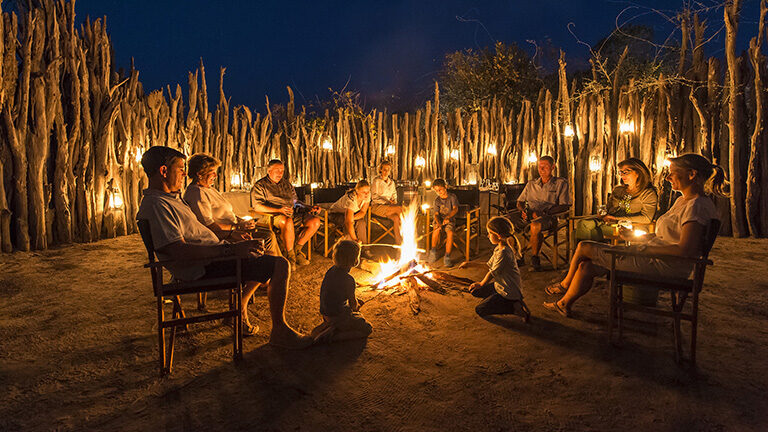 People sitting in a circle around a campfire surrounded by a tall wooden fence at night.