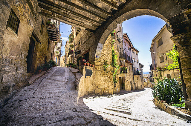 Steep stone street passing under a dark wooden arch into a sunny town plaza. Explore on luxury Spain vacations.