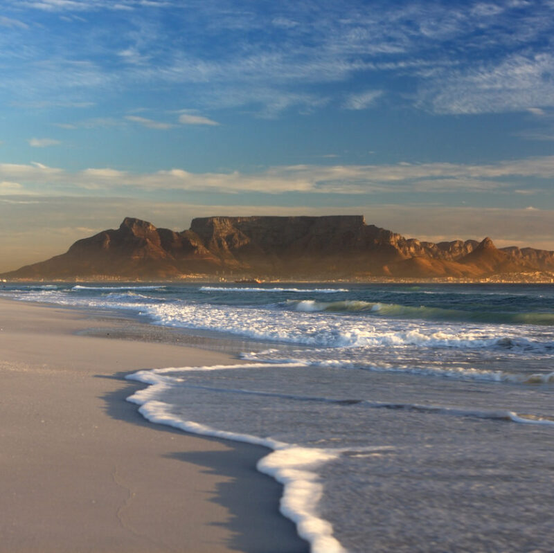 Table Mountain looms over a sandy beach with white sea foam washing ashore under a blue and cloudy sky.