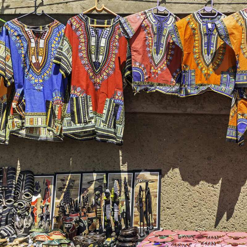 Brightly colored dashiki shirts and various handcrafted African souvenirs displayed for sale at an outdoor market stall.