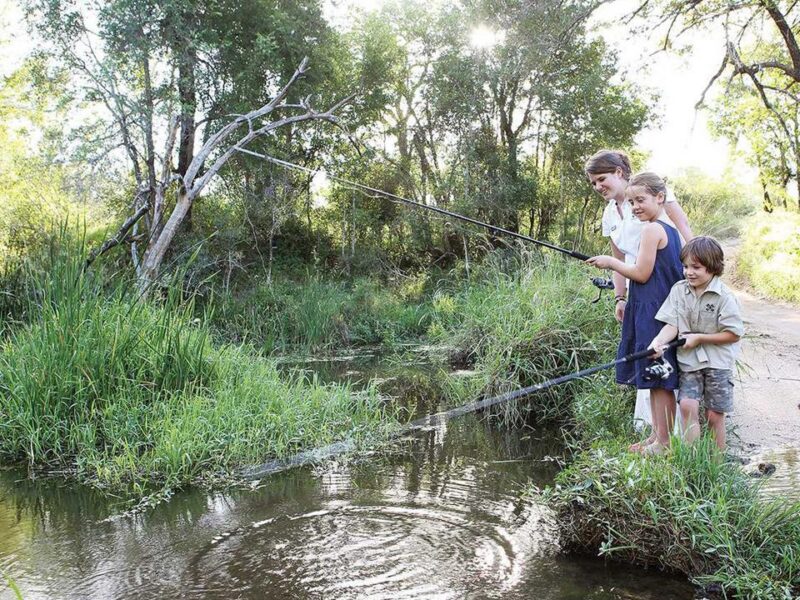 A woman and two children fishing on the grassy bank of a wooded stream during a sunny day.