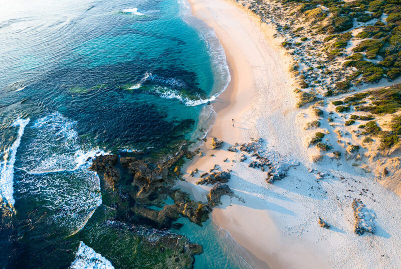 Aerial view of a rugged, curved coastline with white sand, turquoise water, and breaking waves, a beautiful stop on luxury Australia trips.
