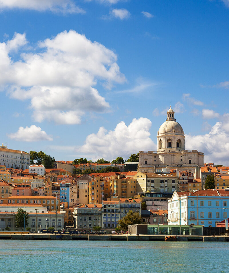 View of Lisbon waterfront with colorful buildings and a large dome church (Santa Engrácia) under a blue sky, luxury Portugal vacations.
