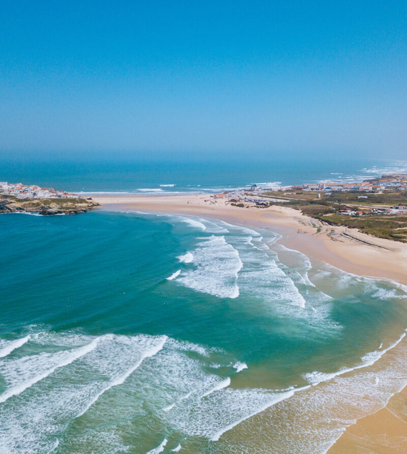 Aerial view of a wide sandy beach, turquoise ocean waves, and a coastal village under a clear sky, ideal for luxury Portugal vacations.