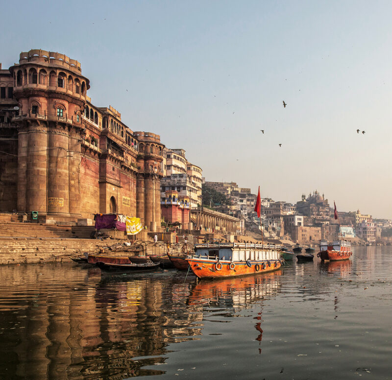 Traditional stone ghats and buildings line the Ganges River, with several boats docked in the foreground in Varanasi on luxury India tours.