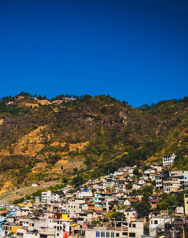Village houses densely packed on a steep, dry hillside with green trees at the top, under a clear, bright blue sky.