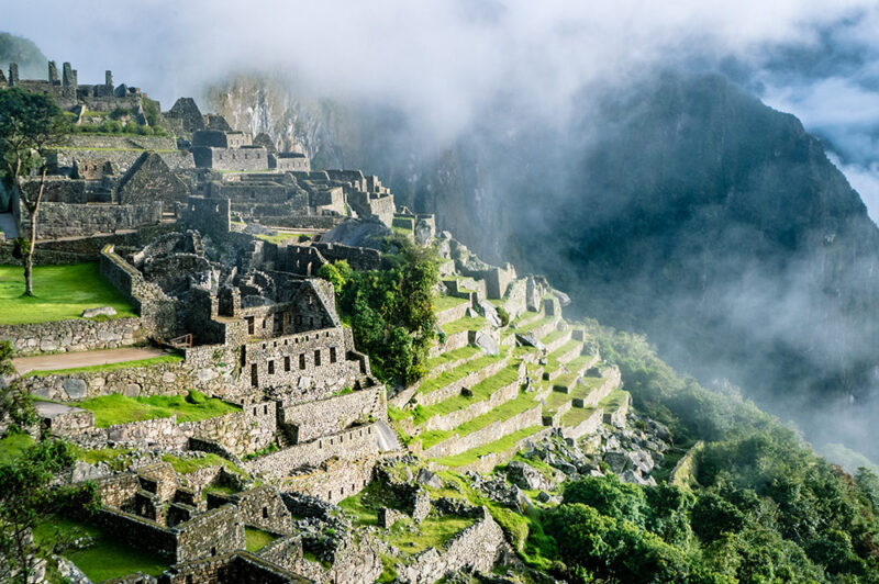 The ruins of Machu Picchu are dramatically visible on a steep, misty mountainside, featuring stone structures and terraces, ideal for luxury Machu Picchu trips.