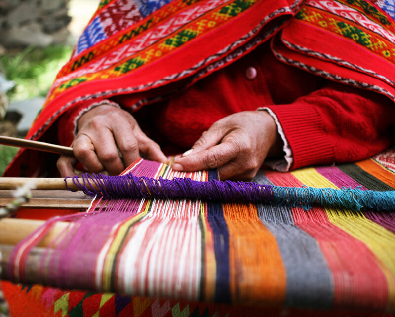 A close-up shows hands weaving a colorful, patterned textile on a wooden loom, with a red cloak draped over the person, ideal for luxury Machu Picchu trips.