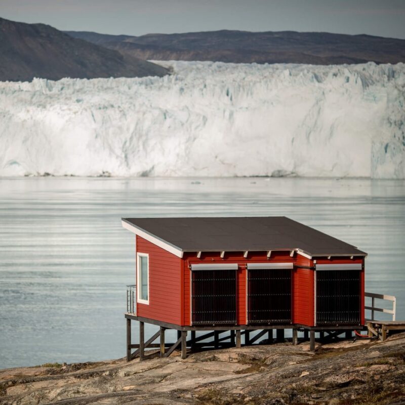 A red wooden cabin on stilts near water and a giant glacier during luxury Polar vacations.