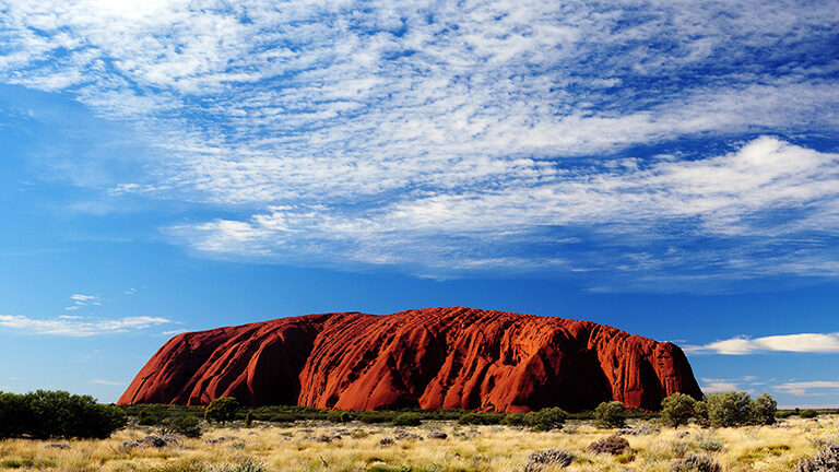 The large red rock formation of Uluru under a bright blue sky for luxury Australasia vacations.