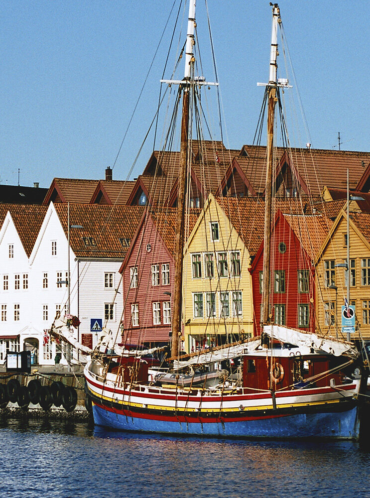 A traditional red, blue, and yellow sailboat docked near historic Bryggen houses in Bergen, Norway, great for luxury Norway tours.