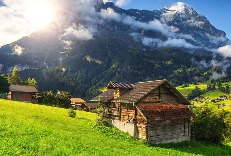 Traditional wooden mountain hut in a sunlit green meadow with snowy peaks for luxury Switzerland vacations.