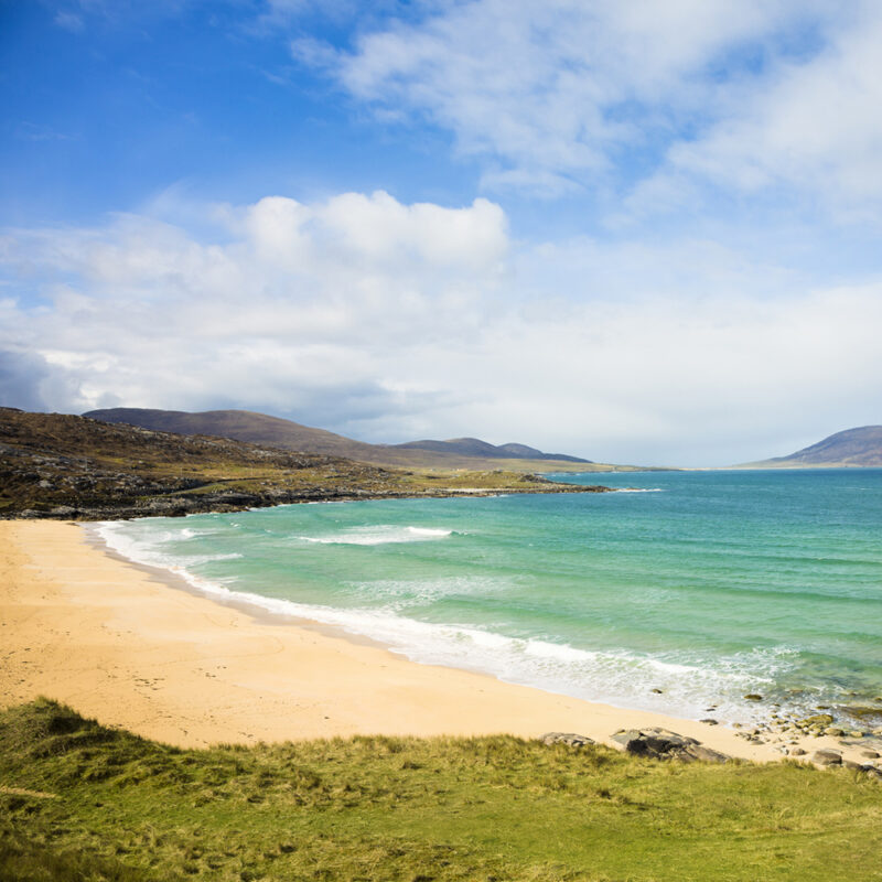 A wide, sandy beach with turquoise water and waves breaking onto the shore, surrounded by grassy hills and blue sky. Ideal for luxury Scotland trips.