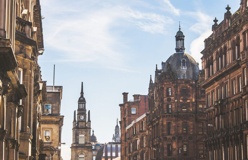 Ornate, historic, red sandstone buildings line a street in Glasgow, with a clock tower and dome against a blue sky. Discover this on luxury Scotland vacations.