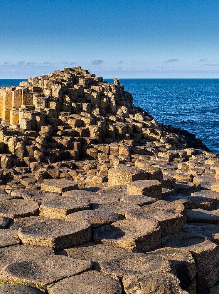 A vertical photo of the Giant's Causeway in Northern Ireland, showing the hexagonal basalt columns meeting the clear blue ocean and sky. Plan your visit on luxury Ireland vacations.
