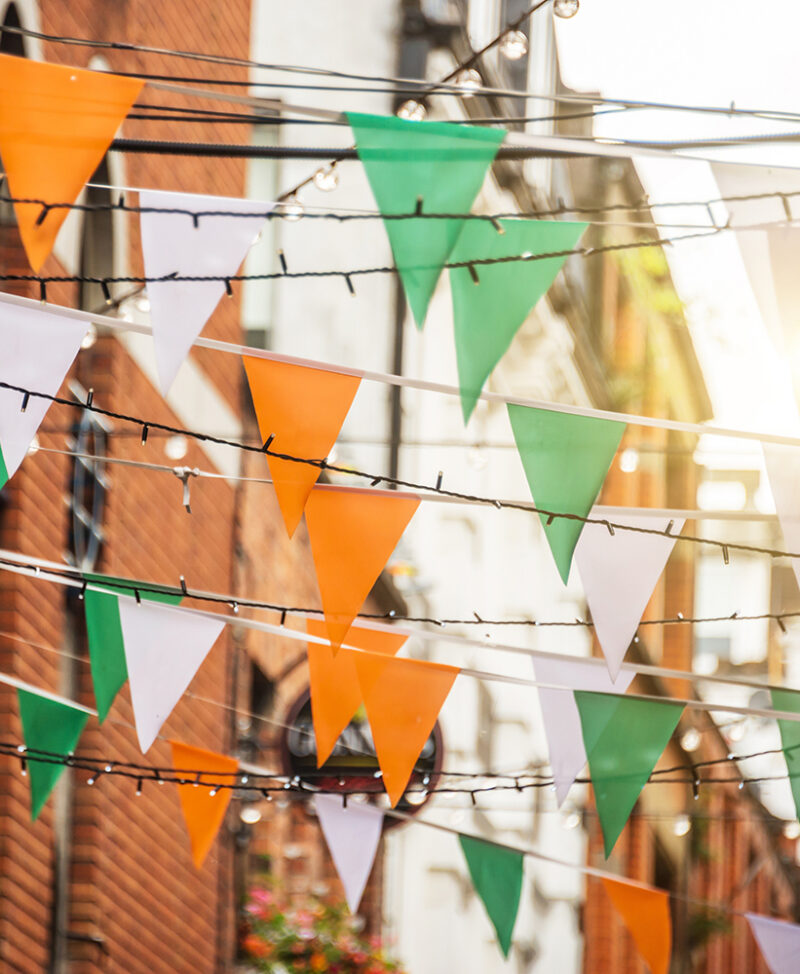 Irish tricolor bunting (orange, white, and green) and fairy lights strung across a street, against a blurred brick building background, offering inspiration for luxury Ireland vacations.