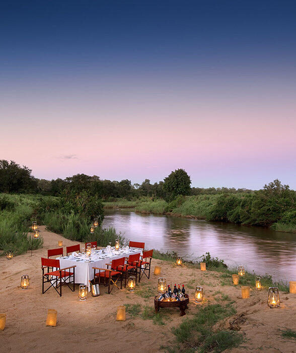 An outdoor dining setup with a long table, red chairs, and many glowing lanterns by a river at dusk.