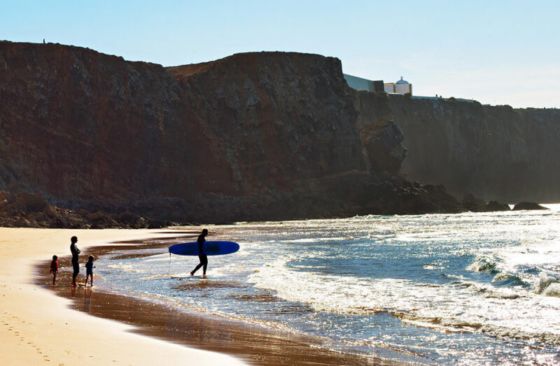 surfer walks by the water's edge under massive sea cliffs during luxury Portugal tours.