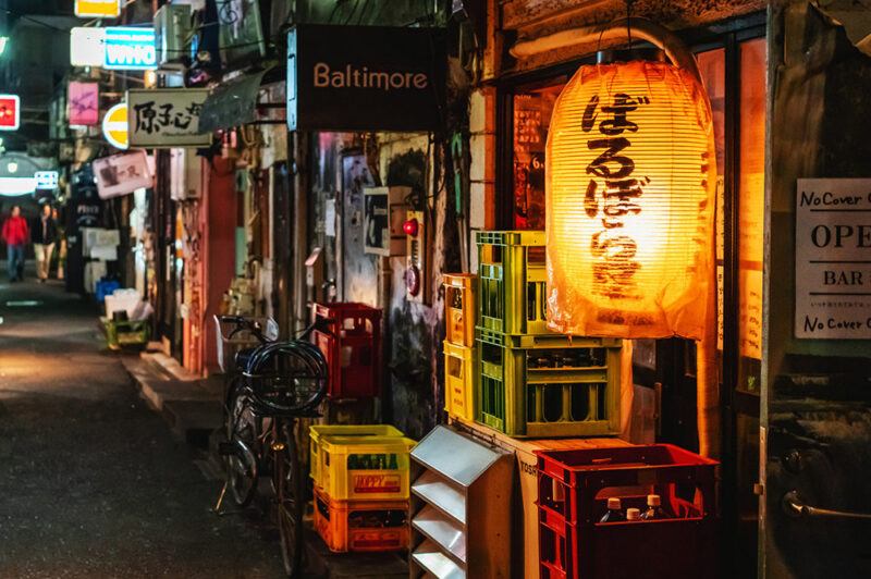 Narrow atmospheric Tokyo alley at night lined with small izakaya bars and illuminated by hanging red lanterns.